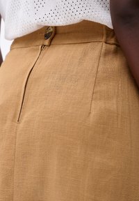 Tan hoge taillebroek gemaakt van textuurstof, met voorste plooien en een enkele knoopsluiting voor een op maat gemaakte look.