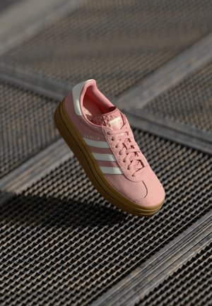 Różowy sneaker Adidas Gazelle z białymi paskami i gumową podeszwą unoszący się nad teksturowaną metalową powierzchnią.