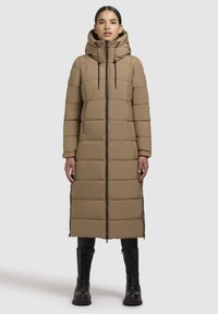 Beige lange gewatteerde jas met capuchon, verticale quiltpatroon, rits aan de voorkant, zijzakken en aangesnoerde manchetten, gedragen met zwarte gevechtslaarzen.