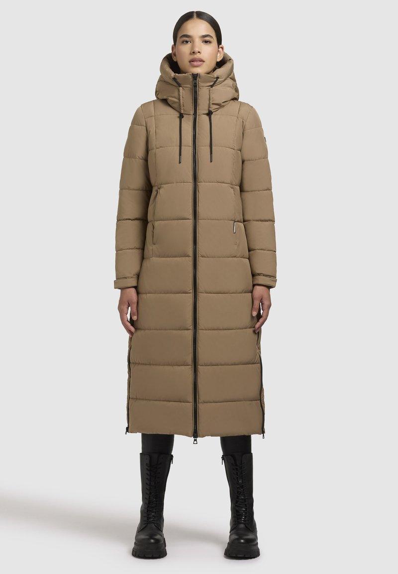 Beige lange gewatteerde jas met capuchon, verticale quiltpatroon, rits aan de voorkant, zijzakken en aangesnoerde manchetten, gedragen met zwarte gevechtslaarzen.