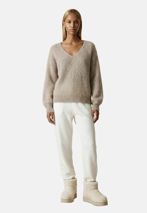 Femme debout face à l'avant, portant un pull beige à col en V, un pantalon blanc et des bottes beige en peau lainée sur fond blanc uni.