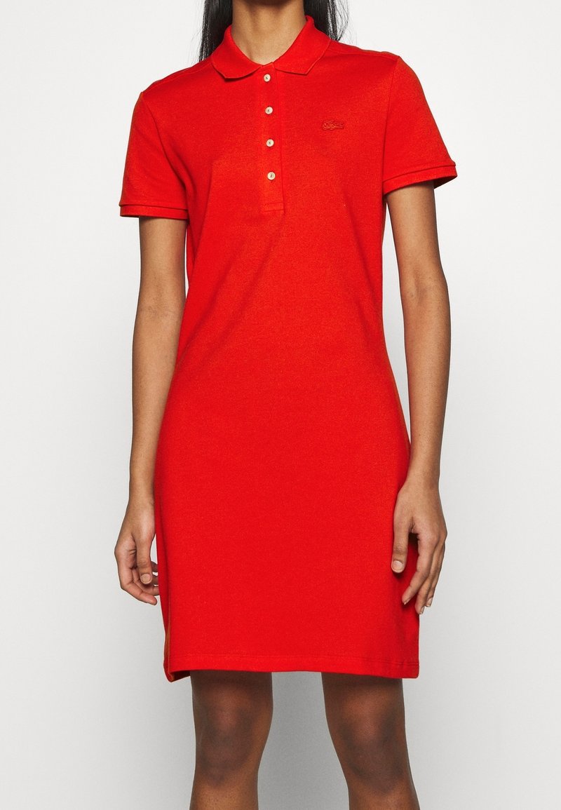 Femme portant une robe polo rouge à manches courtes, longueur genou, avec des boutons et un petit logo sur la poitrine, debout devant un fond uni.