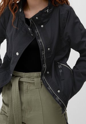 Giacca leggera nera con chiusura a zip, bottoni a pressione e tasche laterali. Presenta cuciture decorative e un design aderente. Pantaloni verde oliva.