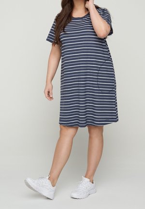 Frau, die ein kurzärmeliges Kleid mit marineblauen und weißen Streifen und weiße Turnschuhe trägt, steht mit einem leicht angehobenen Bein.