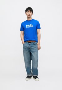 Zils kokvilnas T-krekls ar baltu "KARL LAGERFELD" logotipu, kopā ar gaiši zilām džinsa biksēm un melnām sporta apaviem. Modelis stāv ar rokām kabatās.