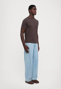 Tricou scurt, de culoare maro, cu gât rotund și un logo mic, purtat alături de blugi despoț de culoare albastră și încălțări maro închis. Design simplu.