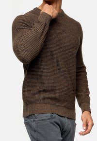 Brązowy sweter z dzianiny o teksturze, z długimi rękawami i okrągłym dekoltem. Noszony z szarymi spodniami, prezentującymi swobodny krój.