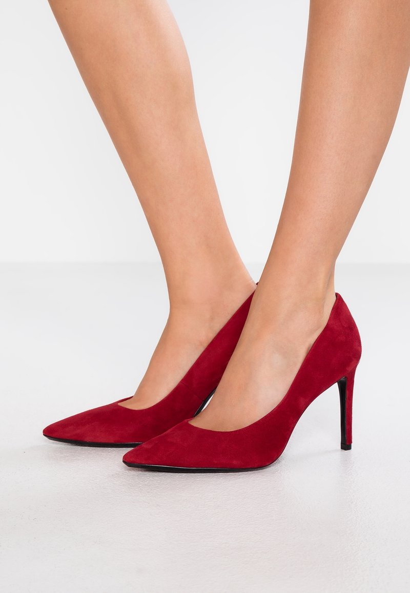 Escarpins hauts en daim rouge avec un bout pointu et des découpes sur le côté ; dotés d'un talon noir élégant. Texture lisse, design minimaliste.