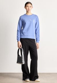 Felpa di cotone azzurro chiaro con logo "GANT", abbinata a pantaloni neri a righe sottili. Tiene in mano una piccola borsa nera lucida con manico arrotondato.