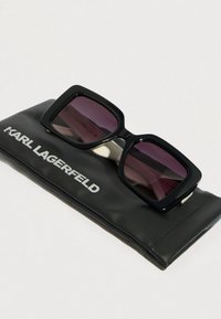 Gafas de sol negras de gran tamaño con lentes degradadas en tono púrpura, colocadas sobre un estuche de cuero negro con la marca blanca "KARL LAGERFELD".