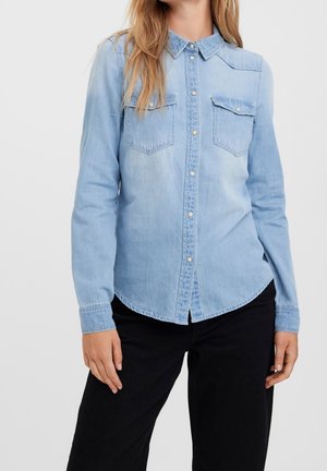 Kvinde iført en lyseblå denimskjorte med knapper og brystlommer samt sorte bukser, stående foran en ensfarvet lys baggrund.