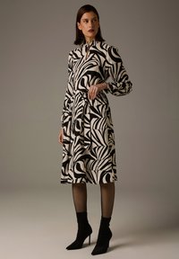 Schwarz-weiß bedrucktes Kleid mit abstrakten, wirbelnden Mustern, langen Ärmeln, hohem Kragen und einem Gürtel zum Binden, kombiniert mit schwarzen knöchelhohen Stiefeln.