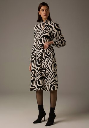 Schwarz-weiß bedrucktes Kleid mit abstrakten, wirbelnden Mustern, langen Ärmeln, hohem Kragen und einem Gürtel zum Binden, kombiniert mit schwarzen knöchelhohen Stiefeln.
