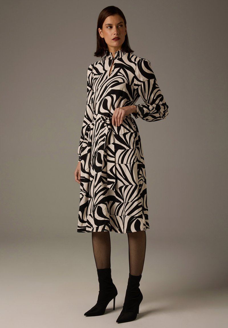 Schwarz-weiß bedrucktes Kleid mit abstrakten, wirbelnden Mustern, langen Ärmeln, hohem Kragen und einem Gürtel zum Binden, kombiniert mit schwarzen knöchelhohen Stiefeln.