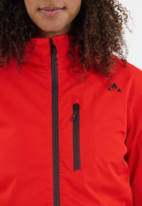 Rood geïsoleerd jack met een hoge kraag, voorzien van zwarte ritsen en een ritssluiting op de borstzak. Gladde textuur, minimalistisch ontwerp.