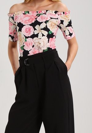 Off-shoulder top met ruches bij de hals, bloemenprint in roze en crème op een zwarte achtergrond; gecombineerd met hoge zwarte broek met een riem.
