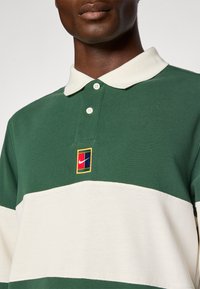 Zielono-kremowa polo z paskami, z klasycznym kołnierzykiem, z małym, kolorowym logo Nike na piersi oraz trzema guzikami.