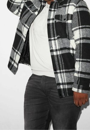 Zwart-wit geruite flanellen jas met een groot ruitpatroon, drukknoopsluiting en een zachte textuur, gedragen over een wit sweatshirt en donkere jeans.