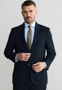 Granatowy garnitur w prążki z klapami z wycięciem, zapięciem na jeden guzik, w połączeniu z jasnoniebieską koszulą i teksturowanym brązowym krawatem.