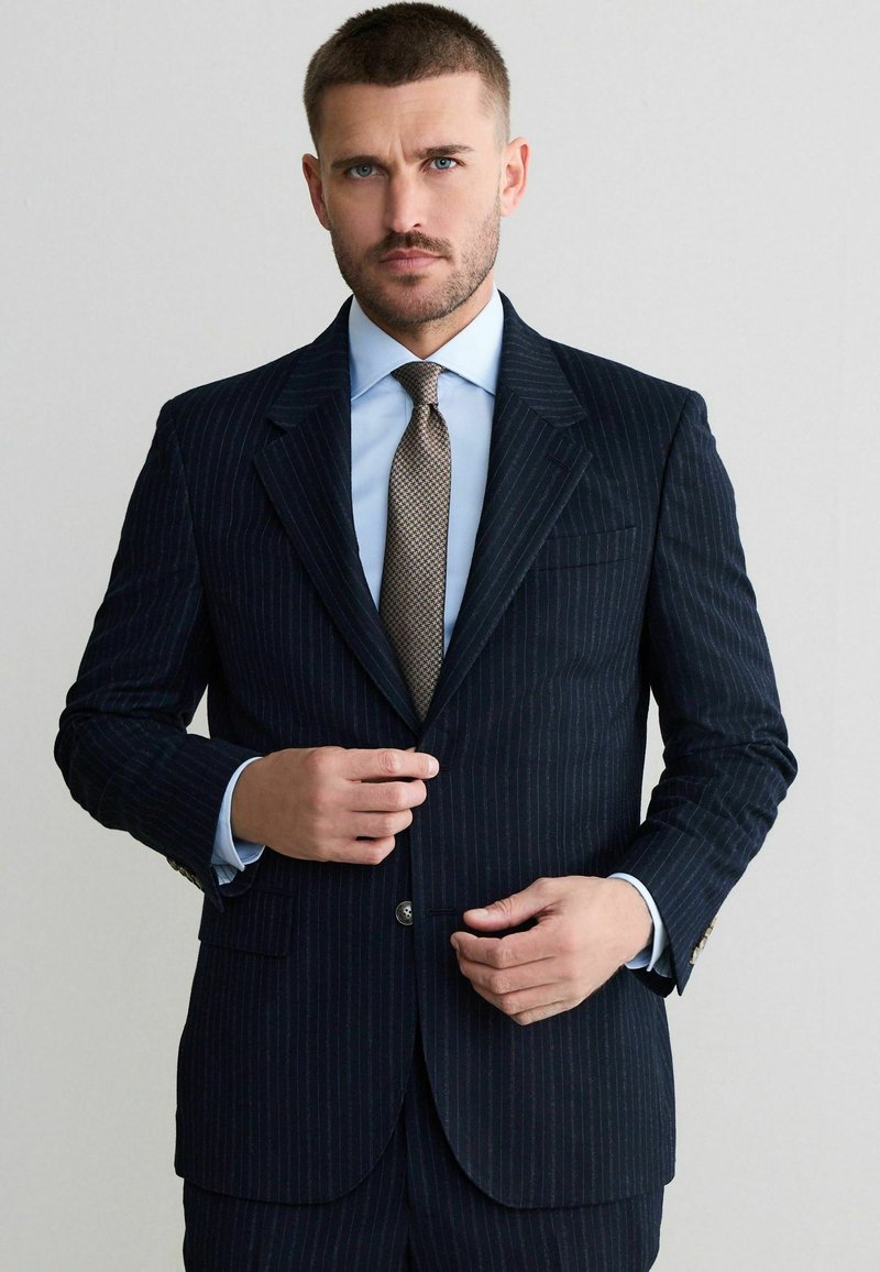 Granatowy garnitur w prążki z klapami z wycięciem, zapięciem na jeden guzik, w połączeniu z jasnoniebieską koszulą i teksturowanym brązowym krawatem.