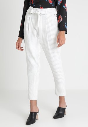 Femme portant un pantalon blanc à fines rayures taille haute avec ceinture, un haut noir à manches longues à fleurs et des chaussures à talons noires sans lacets.