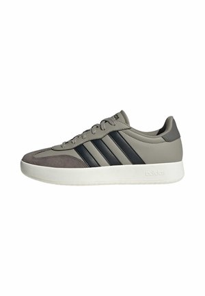 Zapatilla deportiva gris de caña baja con rayas negras, suela blanca, diseño con cordones y puntera de ante, vista lateral.