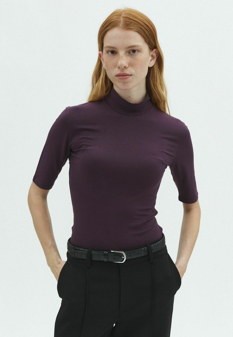 Jeune femme aux cheveux roux portant un col roulé violet ajusté à manches courtes et un pantalon noir avec une ceinture noire texturée, debout les mains dans les poches.