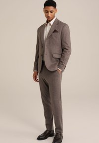 Bruin gestructureerd pak met een bijpassende op maat gemaakte blazer en broek. Inclusief een wit overhemd en donkere gepolijste schoenen. Opmerkelijke details bij de borstzak.