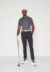 Conjunto de golfe com uma camisa polo cinza, calças pretas, cinto branco, sapatos brancos com detalhes azuis e um boné branco. Segurando um taco de golfe.