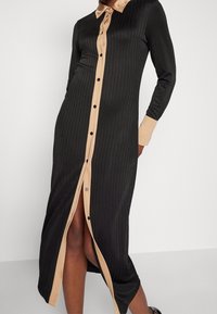 Abito camicia lungo con righe verticali nere, colletto beige a contrasto, chiusura a bottoni e uno spacco a lunghezza media. Tessuto dalla texture liscia.