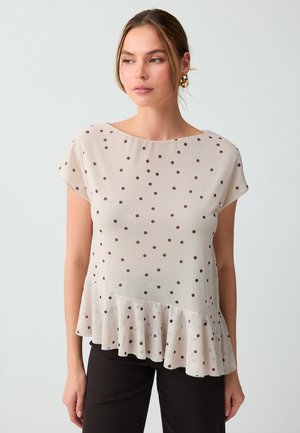 Vrouw draagt een beige blouse met korte mouwen, bruine polkadots en een ruches zoom, gecombineerd met zwarte broek, tegen een effen achtergrond.