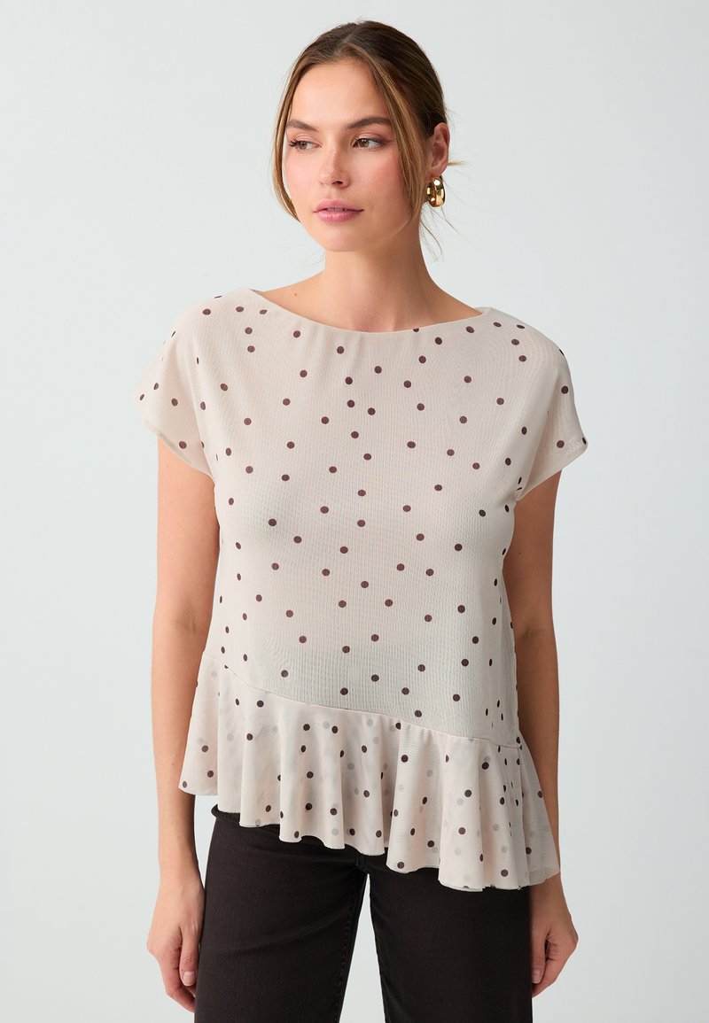 Femme portant un chemisier beige à manches courtes avec des pois marron et un ourlet volanté, associé à un pantalon noir, sur un fond uni.