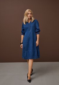 Žena s blond vlasmi, oblečená v modrých rifľových šatách po kolená a čiernych vysokých opätkoch, stojaca pred hnedo-šedým pozadím.