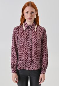 Blusa a maniche lunghe in un profondo bordeaux con un motivo geometrico bianco. Presenta un colletto classico con dettagli beige e chiusura frontale con bottoni.