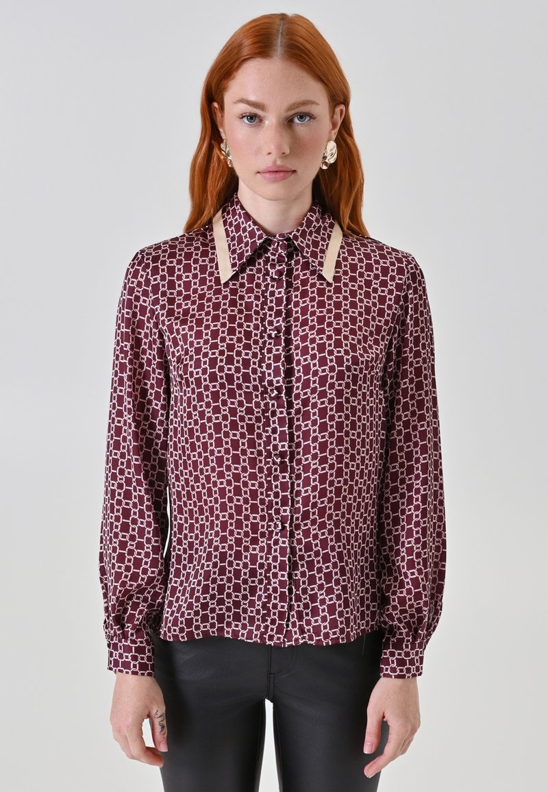 Blusa a maniche lunghe in un profondo bordeaux con un motivo geometrico bianco. Presenta un colletto classico con dettagli beige e chiusura frontale con bottoni.