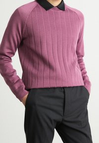 Personne portant un pull violet à côtes à manches longues par-dessus une chemise noire à col, associée à un pantalon noir, mains dans les poches.