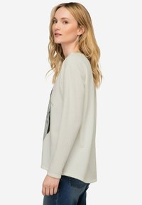 Camisetta a maniche lunghe di colore crema con vestibilità ampia, caratterizzata da una stampa grafica, scollatura rotonda e tessuto testurizzato. Abbinata a jeans blu.