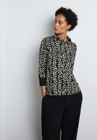 Czarny i kremowy top w wzory z dekoltem typu carmen oraz trzema ćwiartkami rękawów, wykonany z miękkiego materiału, w zestawieniu z luźnymi ciemnymi spodniami.