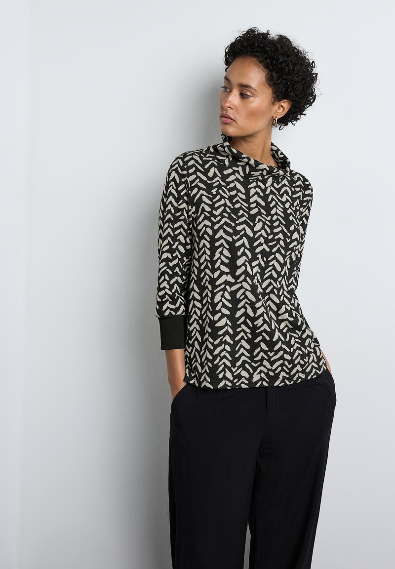 Czarny i kremowy top w wzory z dekoltem typu carmen oraz trzema ćwiartkami rękawów, wykonany z miękkiego materiału, w zestawieniu z luźnymi ciemnymi spodniami.