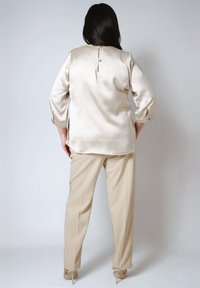 Woman standing with back facing camera wearing beige satin blouse, matching beige pants, and beige high-heeled shoes against plain background.