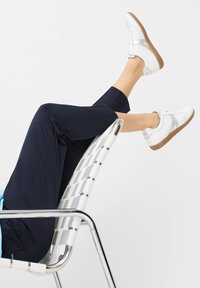 Weiße Sneakers mit silbernen Akzenten, kombiniert mit navyfarbener nicht ganz langer Hose und auf einem silbernen, geflochtenen Stuhl ruhend. Klare Linien und lässiges Design.
