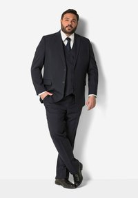 Dunkelblaues Dreiteiliges Anzug mit Reverskragen, einreihiger Jacke und Knopfdetails. Kombiniert mit einem weißen Hemd und einer dunklen Krawatte.