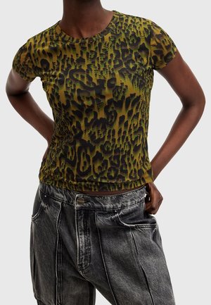 Persona con una camiseta de manga corta con estampado de leopardo en verde y negro, con vaqueros de mezclilla negros desteñidos y de tiro alto, con las manos apoyadas en las caderas.