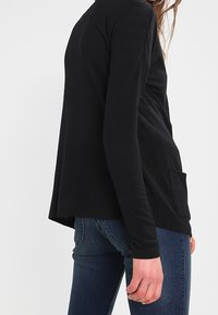 Kobieta w czarnej bluzce z długim rękawem i kieszenią po boku oraz ciemnoniebieskich dżinsach, widziana z boku na jednolitym tle.