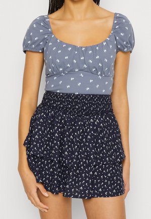 Graues Oberteil mit Puffärmeln und Blumenmuster sowie einem marineblauen, rüschierten Rock mit einem ähnlichen Blumendesign und einem gerafften Taillenbund.