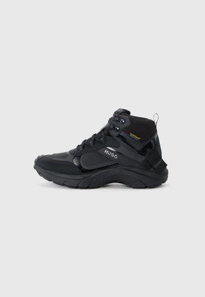 Zapatilla negra de media altura con material texturizado, puntera redonda, diseño de cordones seguro y branding lateral. Cuenta con una suela acolchada y un agarre fuerte.