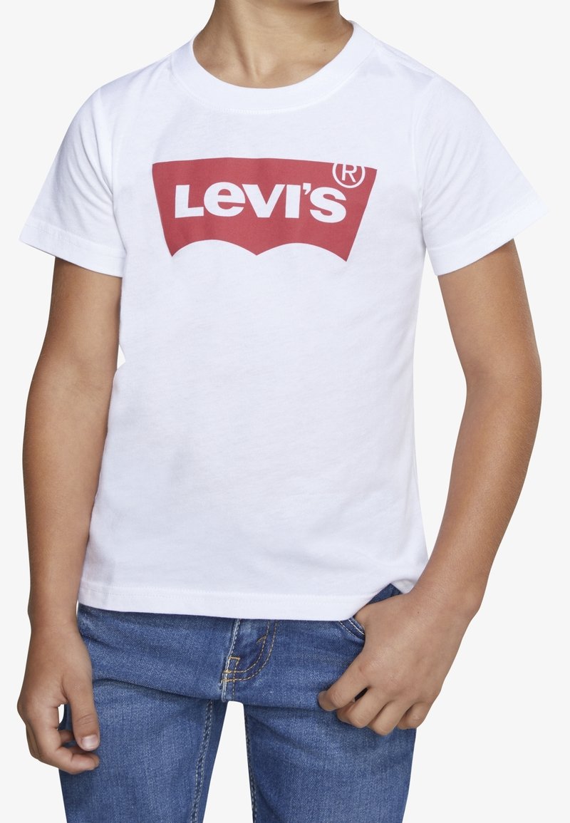 Enfant portant un t-shirt blanc Levi’s avec un logo rouge et un jean bleu en denim, debout, une main dans la poche et l’autre détendue.