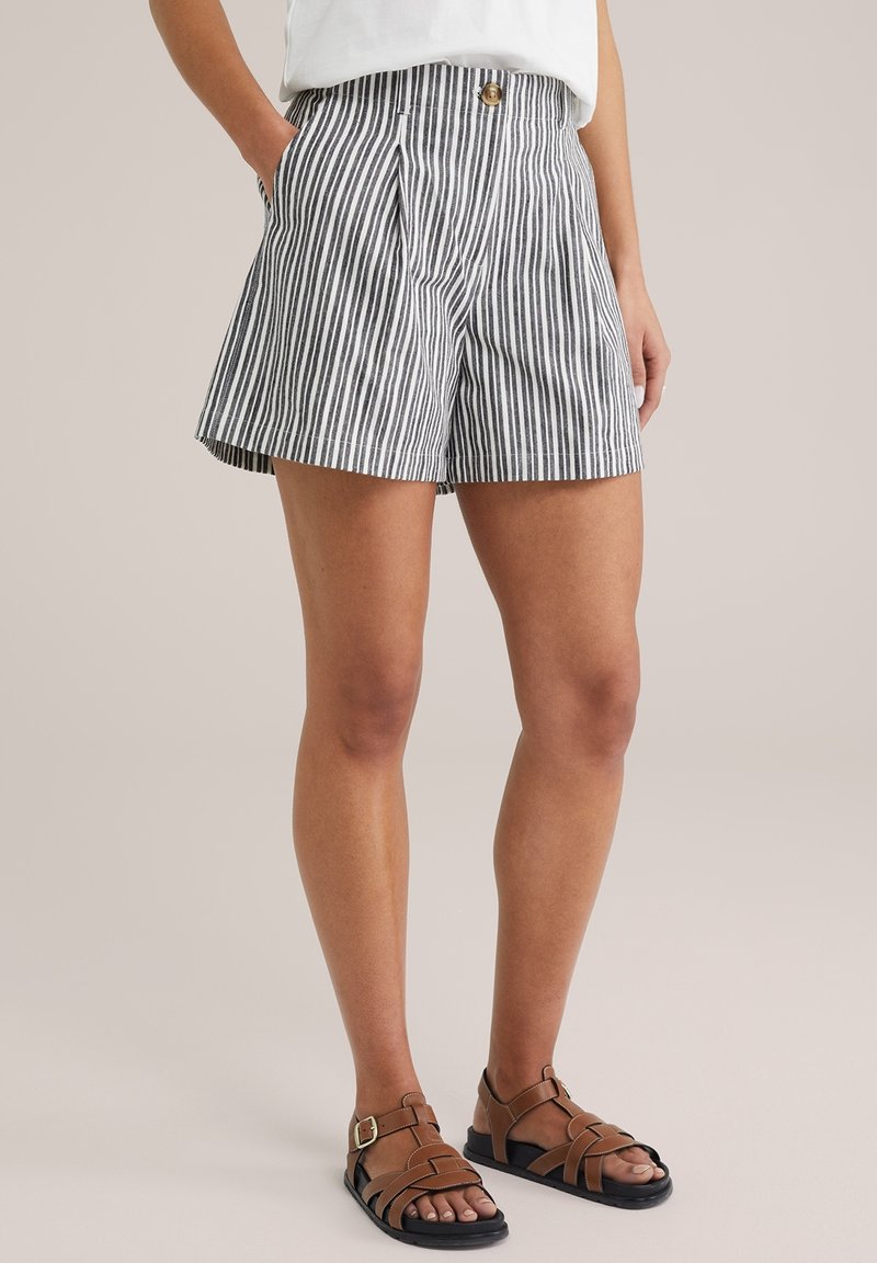 Donna che indossa pantaloni corti a righe verticali bianchi e neri, una maglietta bianca e sandali con fibbia marroni su uno sfondo uniforme.