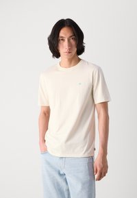 Kurzärmliges cremefarbenes T-Shirt aus weichem Material, versehen mit einem kleinen hellblauen Logo auf der Brust, kombiniert mit hellblauen Denim-Jeans.