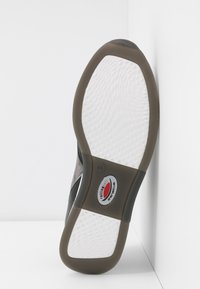 Sulan på skon har en vit texturerad gummiyta med grå accenter. En logotyp är synlig nära mitten och hälen har en svart kant.